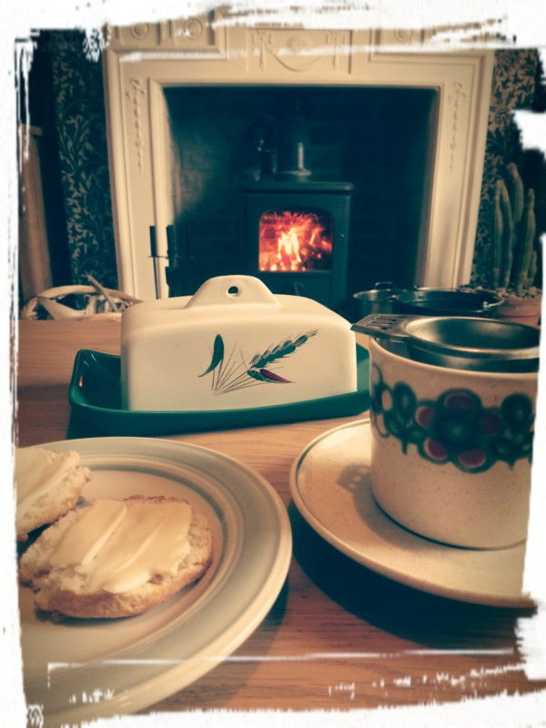 Unpackaged Food Revolution - image of a cup with tea strainer for straining loose leaf tea from the pot, butter dish and plate of buttered scones, with wood burner alight in the background - tea by the fire.